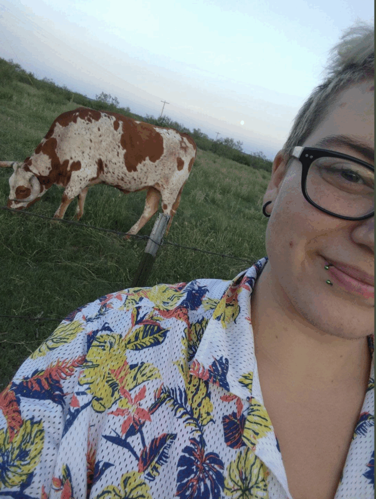 Wichahpi King in front of a cow. Wichahpi is half-cut off since it's a selfie. They wear a printed shirt with a white background and multi-colored trees on it. In the background is a field with a brown and white spotted cow grazing.